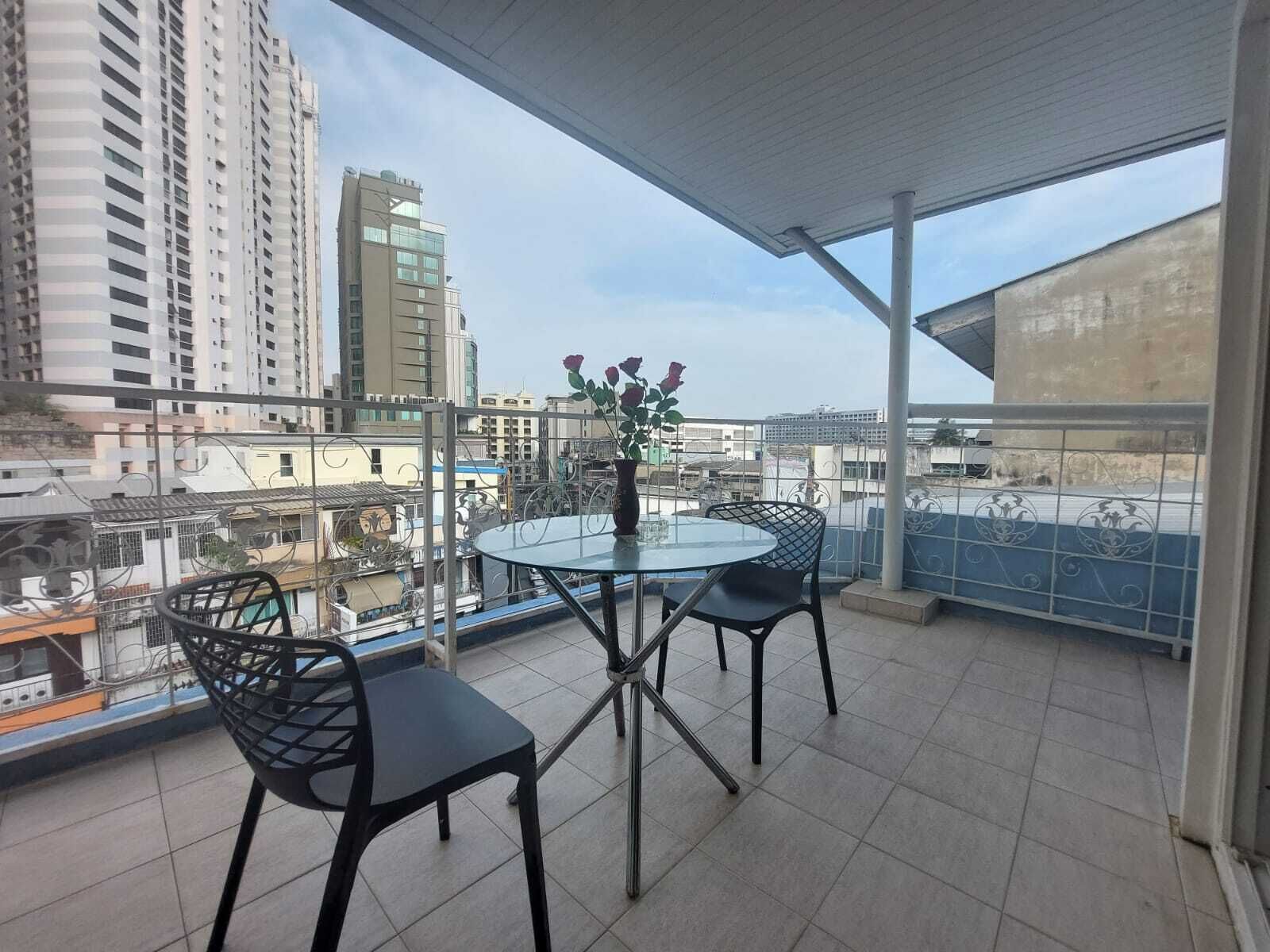 A balcony with a glass table and two chairs, a vase with flowers, and a cityscape view.