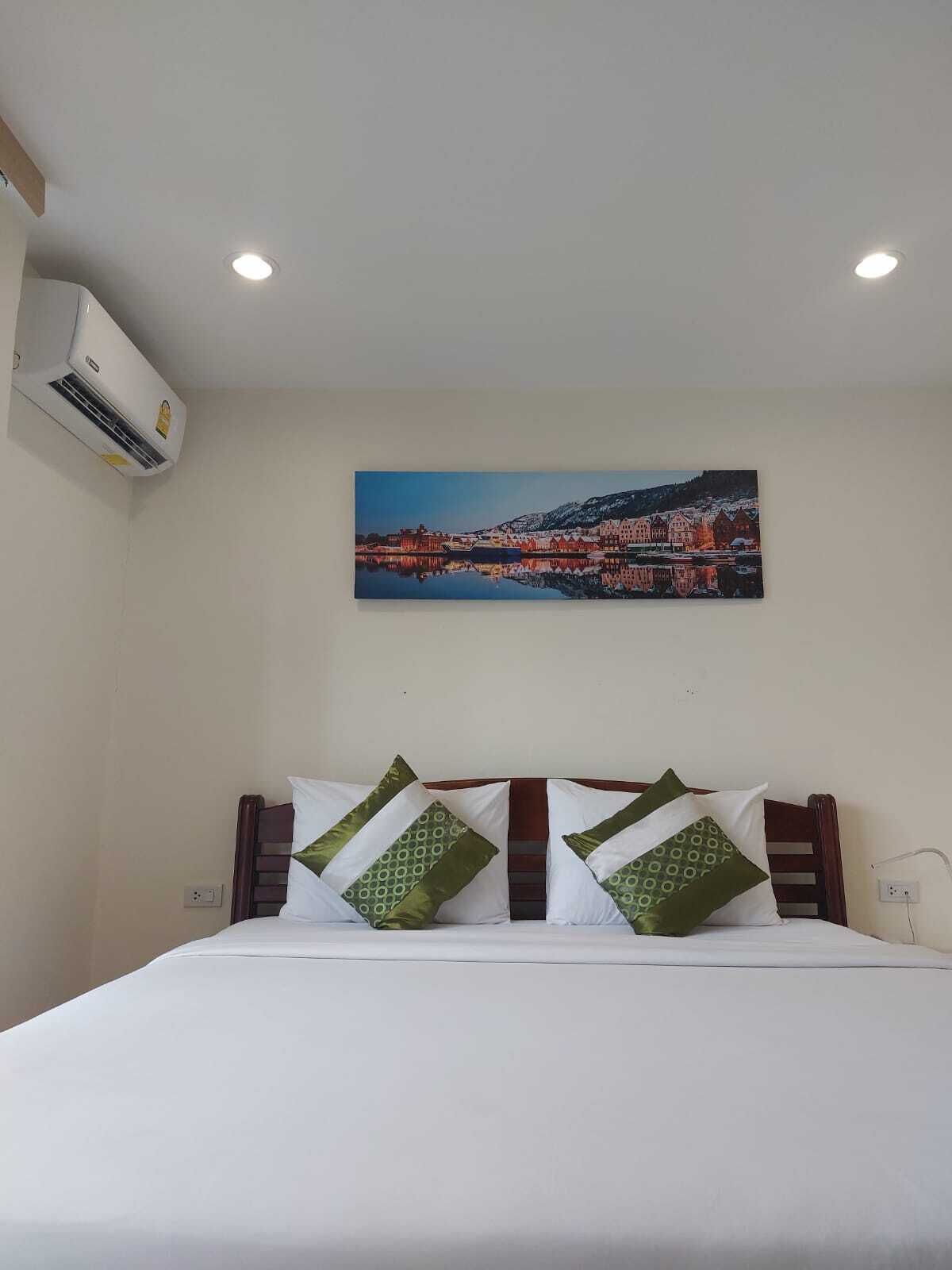 A hotel room with a bed, pillows, an air conditioning unit, and a cityscape painting on the wall.