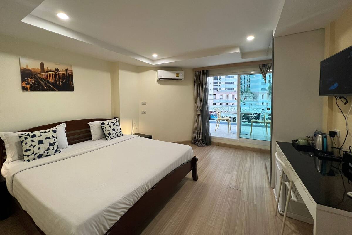 A hotel room with a large bed, a desk, a TV, and a balcony with a view of buildings.