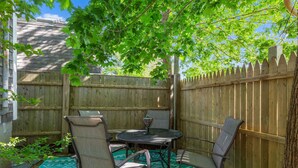 En terrasse med et rundt bord og fire stole, omgivet af et træhegn og et træ der giver skygge.