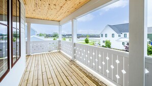 Une véranda couverte, dotée d’un plancher en bois et de rampes blanches, qui offre une vue sur un quartier résidentiel avec ses maisons et un pont.