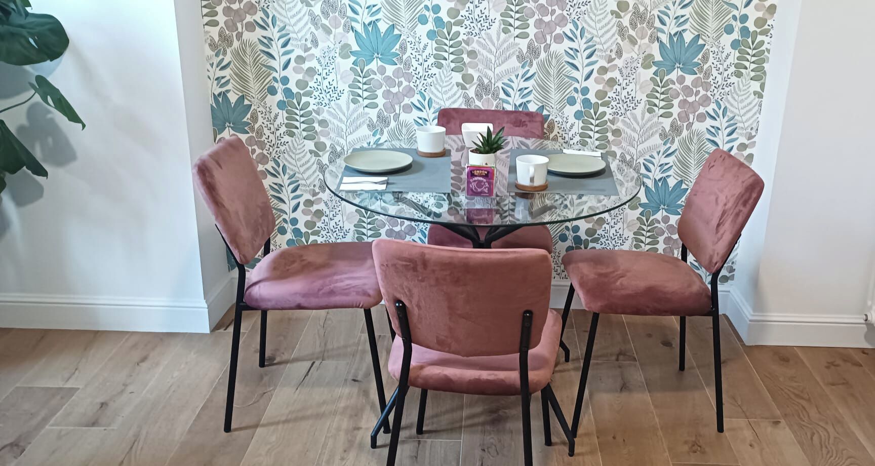 A dining table with four chairs, a glass top, and a floral wallpaper background.