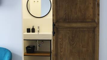 A bathroom with a wooden door, a round mirror, a sink, and a blue chair.