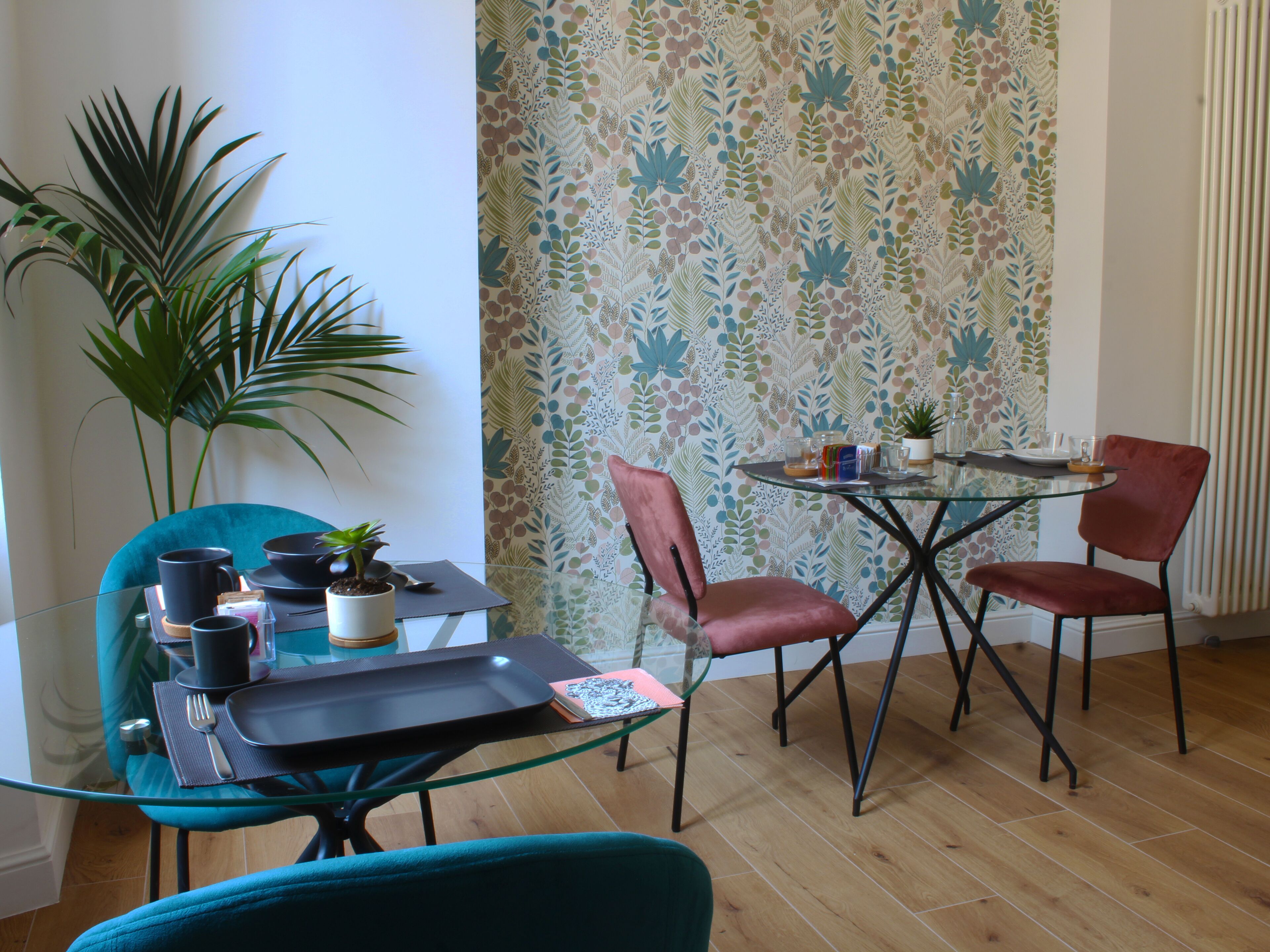 A dining area with a glass table, two chairs, a potted plant, and a patterned wallpaper.