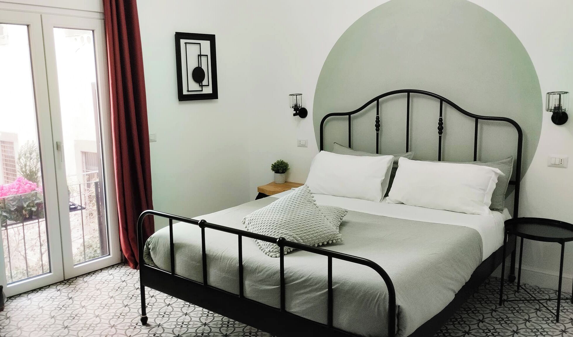 Un letto con struttura in metallo, un piccolo comodino con una pianta, una lampada a parete e vista attraverso una porta in vetro.