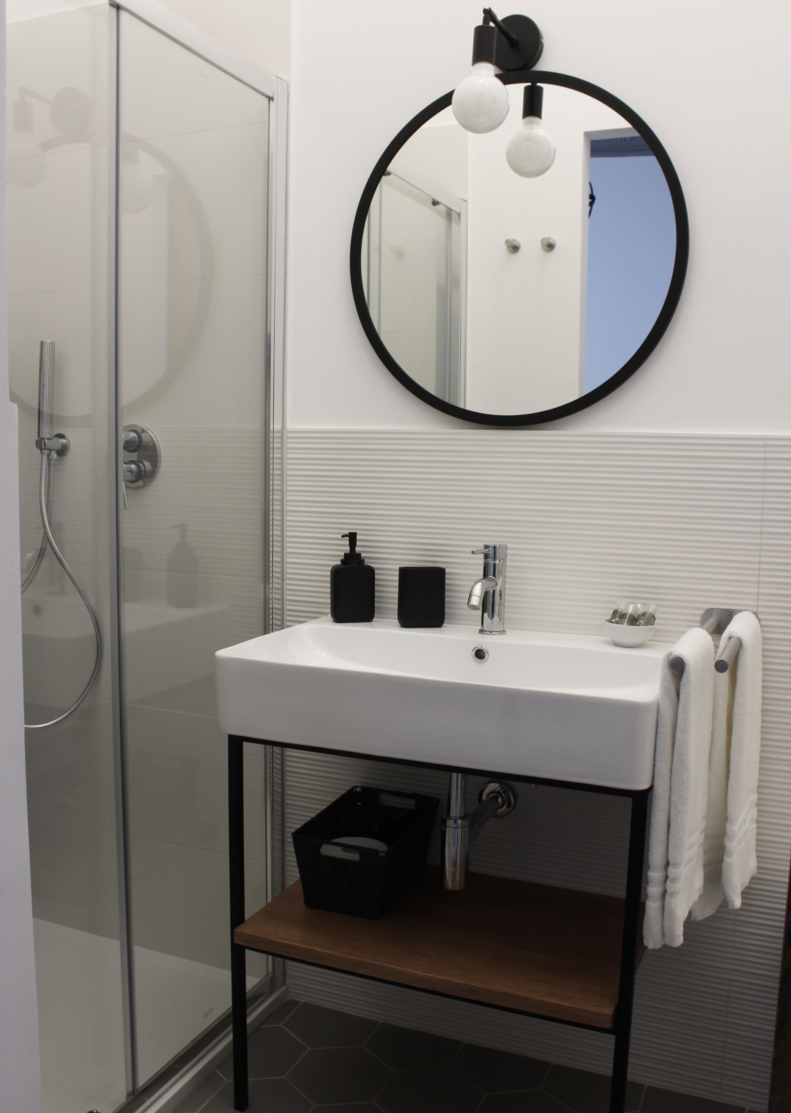 A bathroom with a glass shower enclosure, a white sink with a black soap dispenser, and a round mirror with two light bulbs.