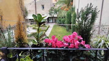 Un balcón con plantas en macetas, vistas a un jardín cuidado y un edificio con tejado de tejas.
