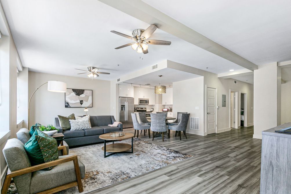 A spacious living area with a gray sofa, a coffee table, and a dining set. There are ceiling fans, a kitchen in the background, and a large window.