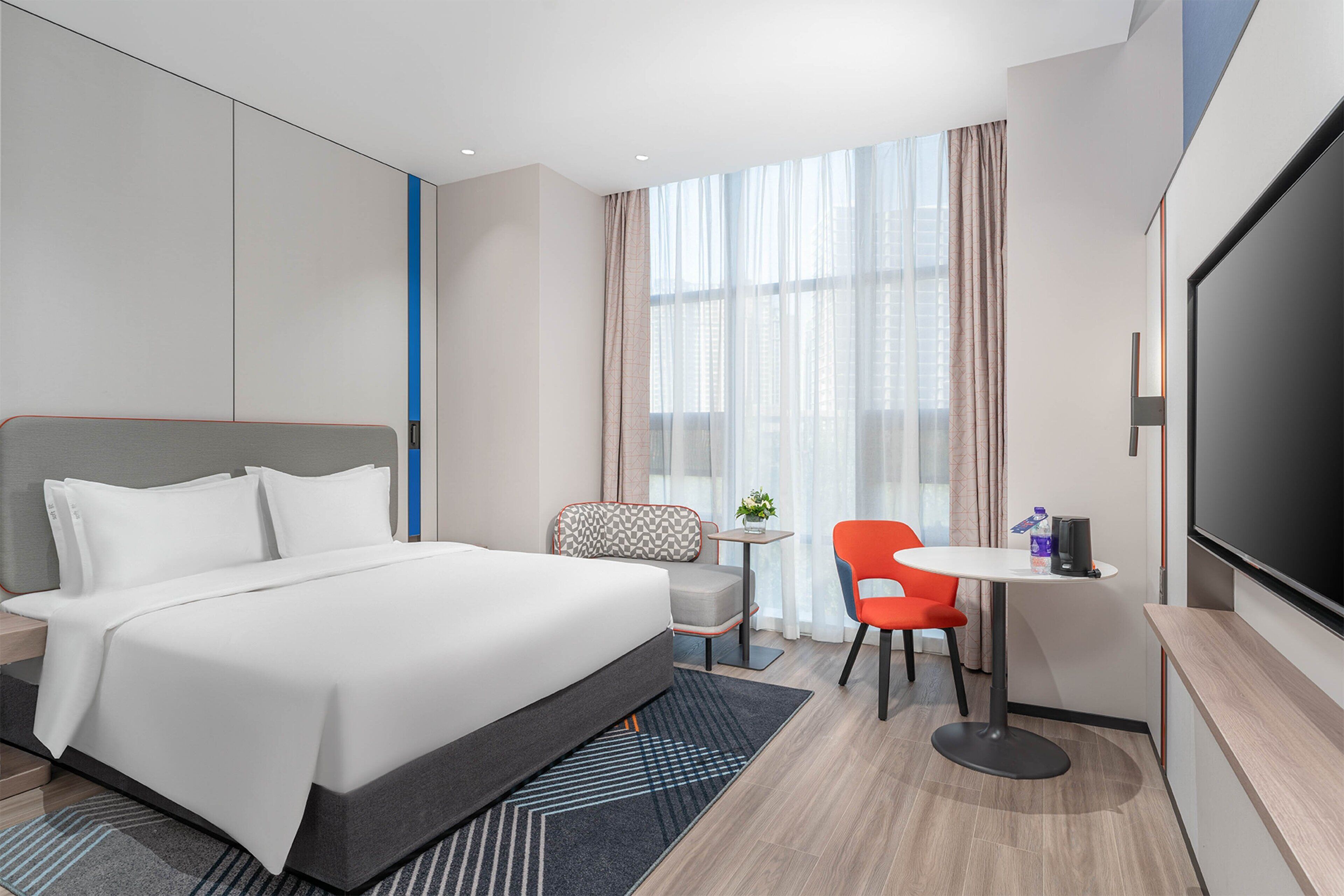 Una habitación de hotel moderna con una cama grande, un escritorio con una silla, un televisor y un ventanal con cortinas.