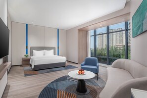 Een moderne hotelkamer met een groot bed, een bank, een bijzettafeltje met fruit en een stadszicht door grote ramen.