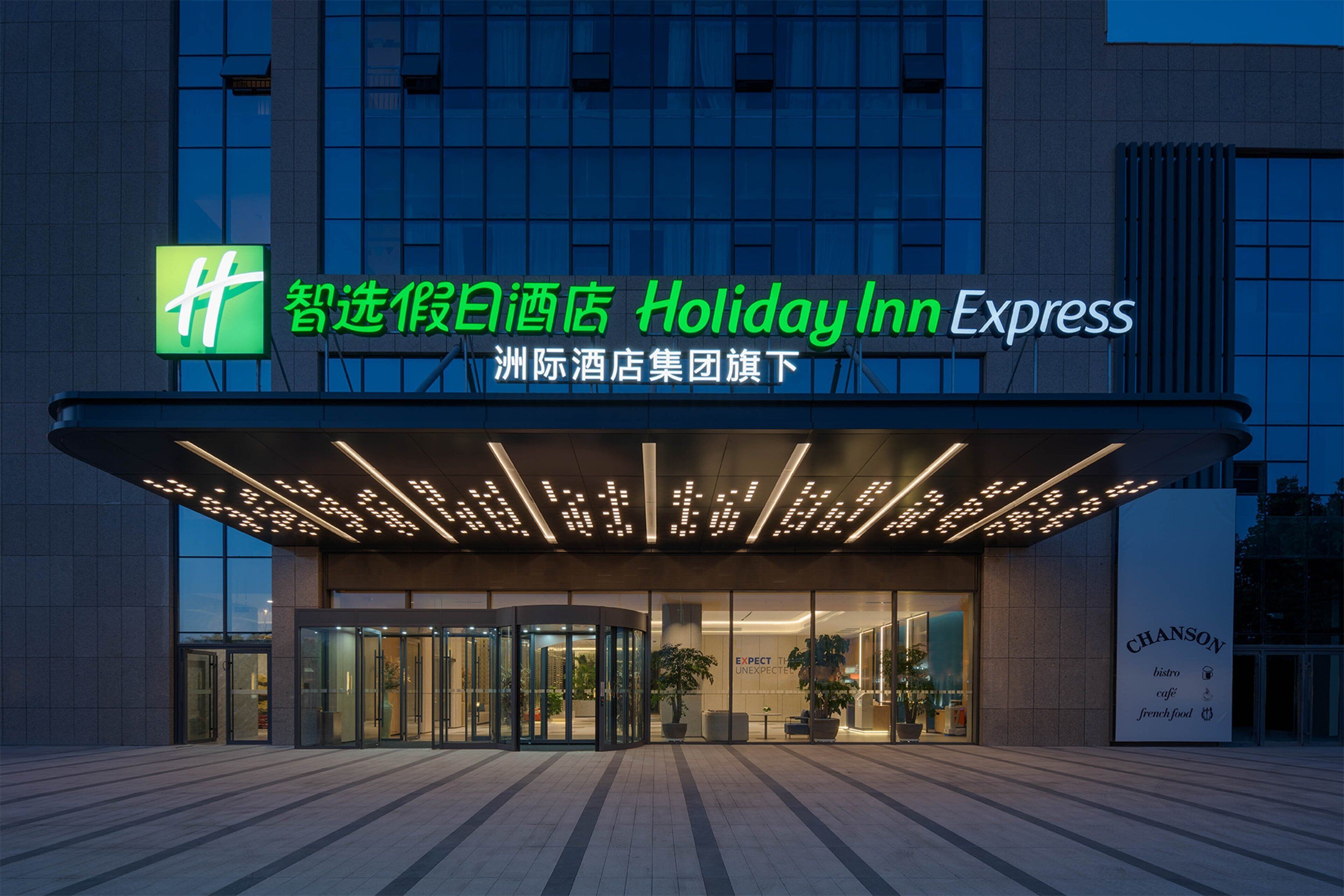 A Holiday Inn Express hotel entrance with a glass facade, automatic revolving doors, and a signboard displaying the hotel name and logo.