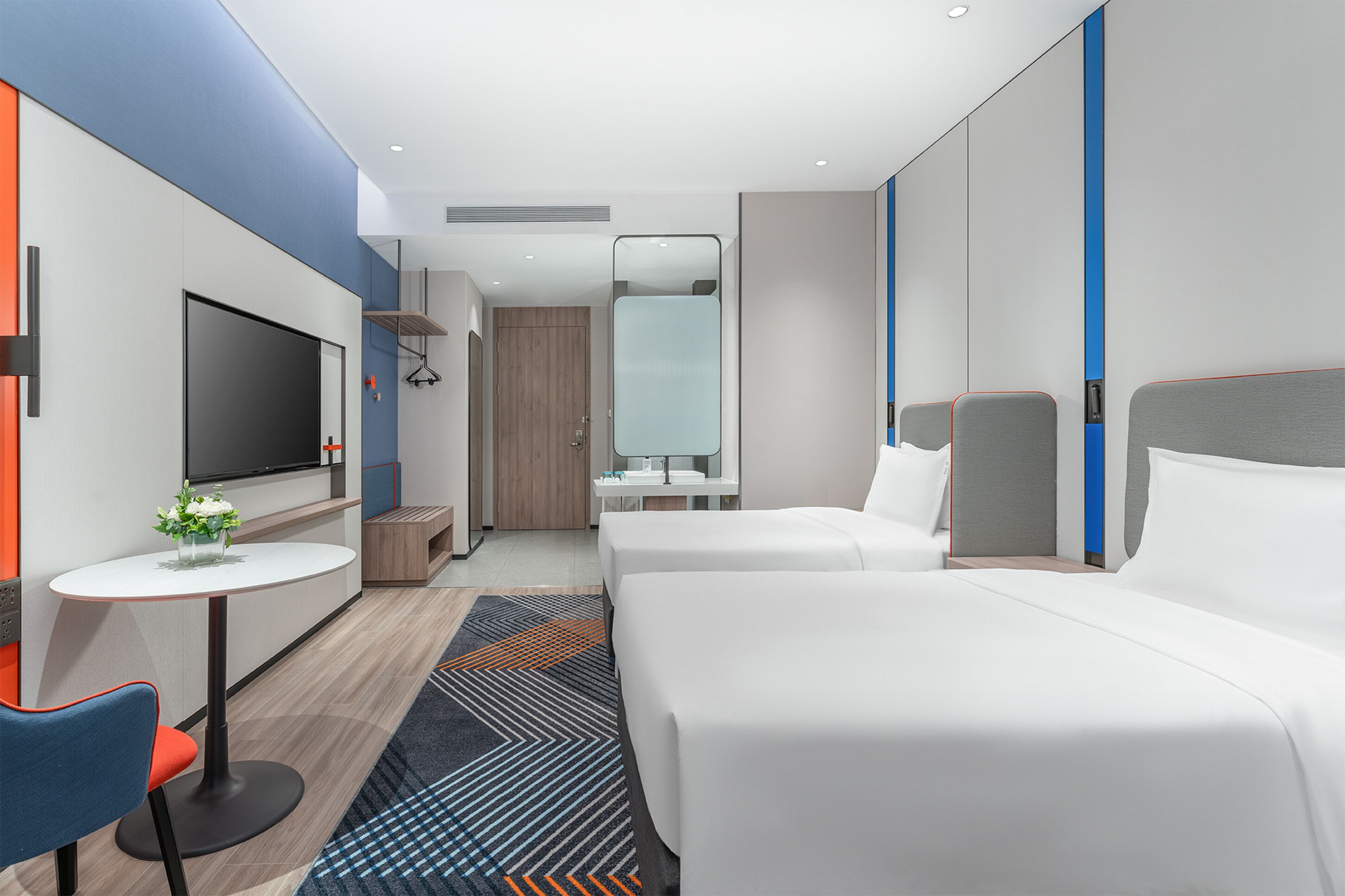 A modern hotel room with a flat-screen TV, a small table with a vase of flowers, two beds, a mirror, and a wardrobe.
