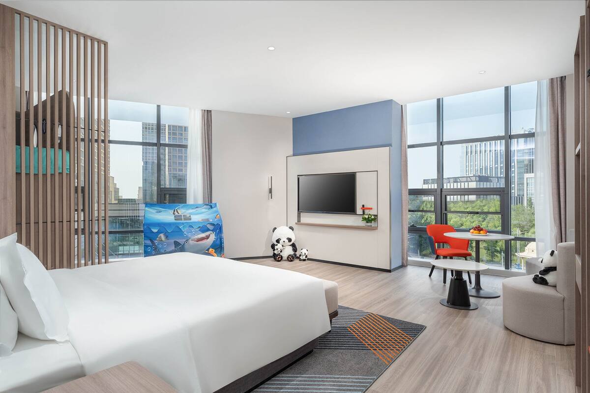 Une chambre d’hôtel moderne dotée d’un grand lit, d’un téléviseur fixé au mur, d’un coin repas avec une table et des chaises, et de grandes fenêtres offrant une vue sur la ville.