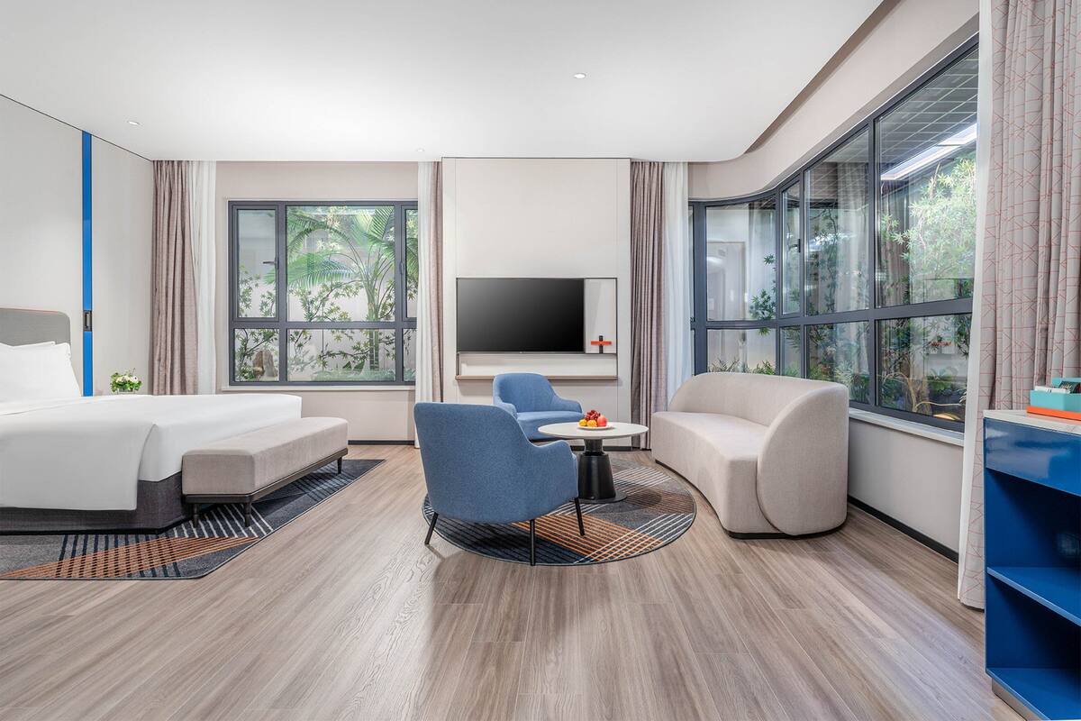 Une chambre d’hôtel moderne équipée d’un lit, de deux fauteuils, d’un canapé, d’une télévision et de grandes fenêtres donnant sur un paysage verdoyant.