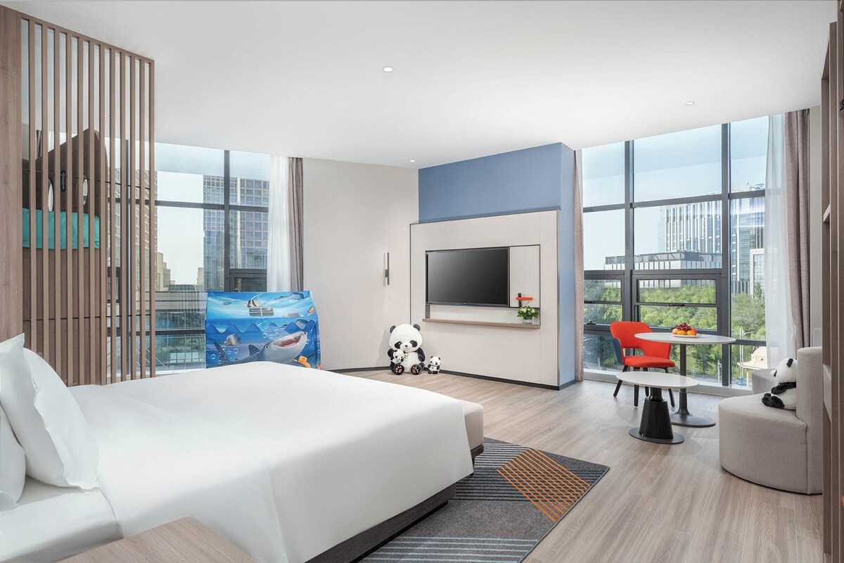Een moderne hotelkamer met een groot bed, een aan de muur bevestigde televisie, een zithoek met een tafel en stoelen, en grote ramen met uitzicht over de stad.