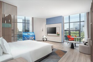 Una habitación de hotel moderna con una cama grande, un televisor montado en la pared, un comedor con mesa y sillas, y ventanas amplias con vistas a la ciudad.
