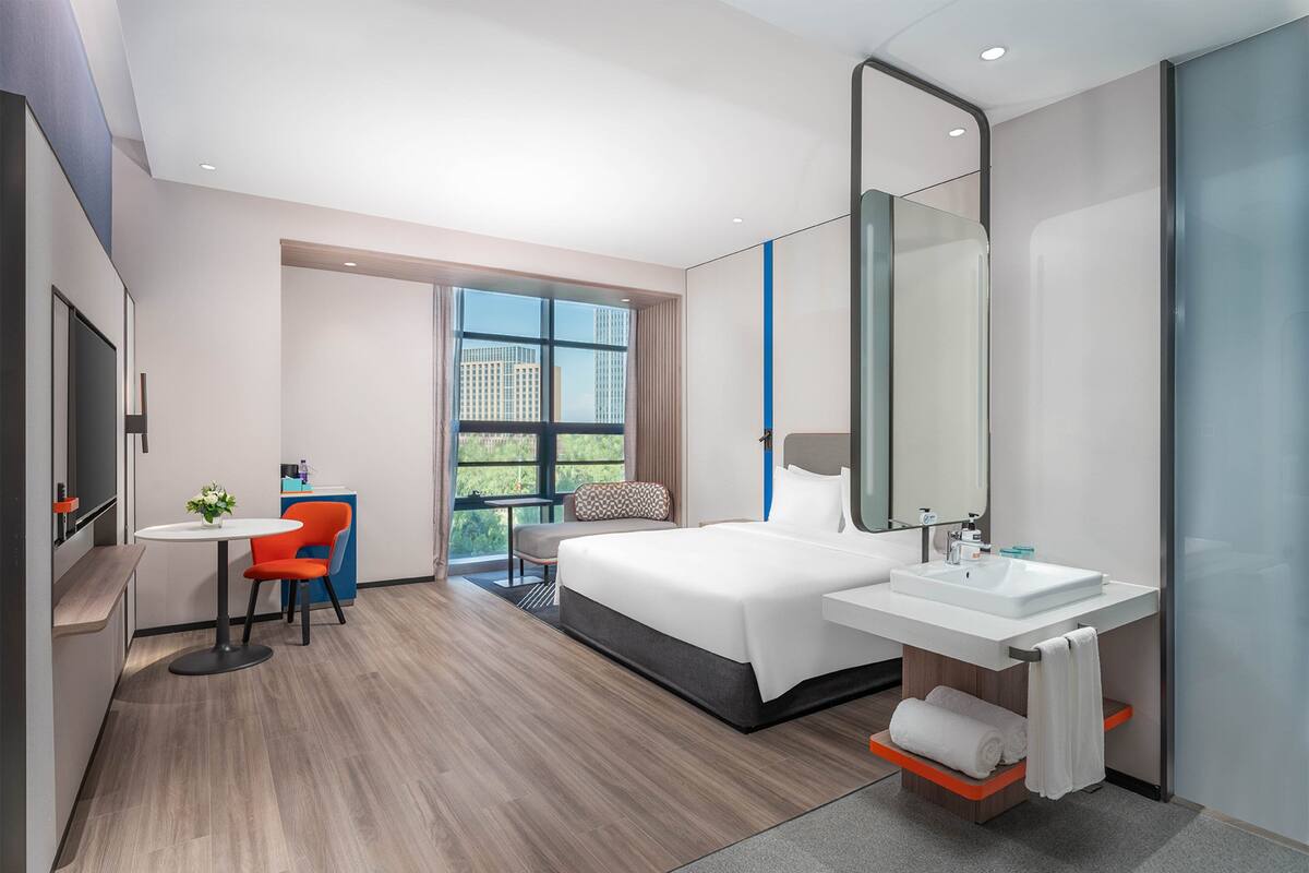 Une chambre d’hôtel moderne dotée d’un grand lit, d’un bureau avec une chaise, d’une salle de bain avec un lavabo et un miroir, et d’une fenêtre donnant sur la ville.
