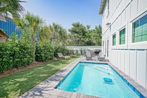 A rectangular swimming pool with a tiled edge, surrounded by a brick patio. There are lounge chairs and a barbecue grill in the background.