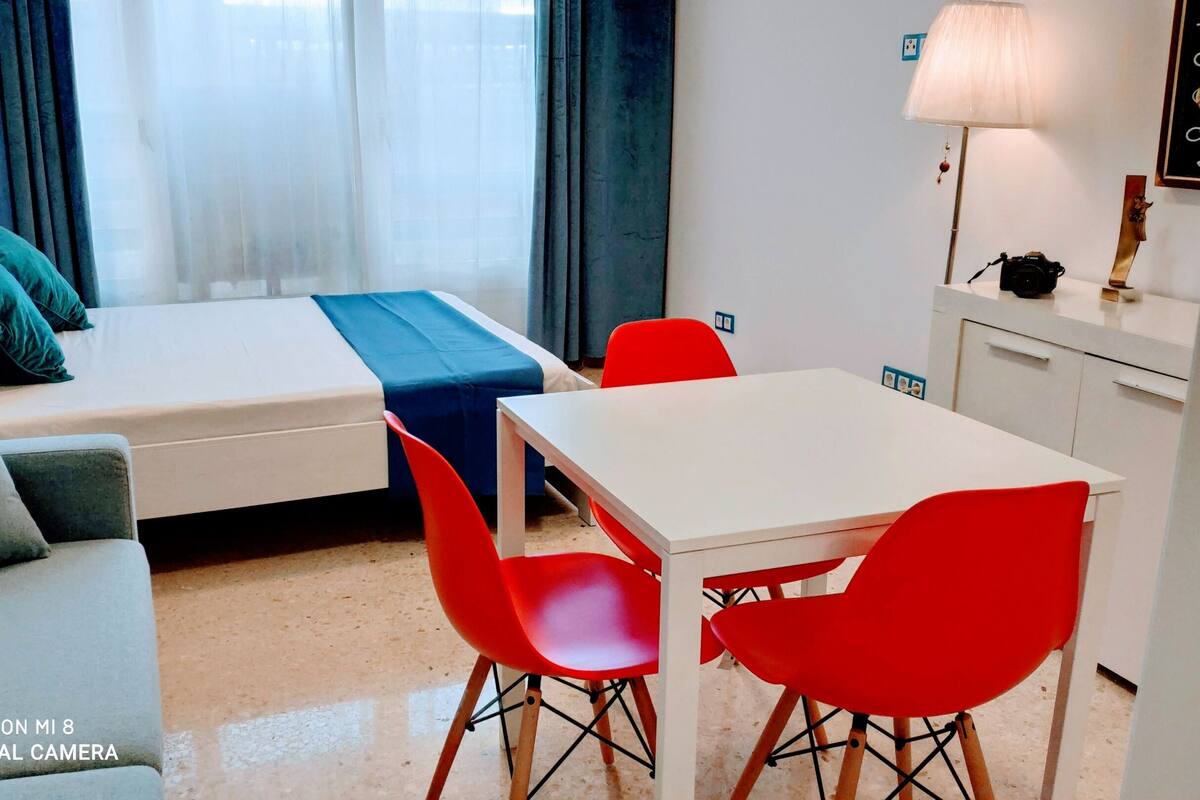A compact hotel room with a bed, a small sofa, a dining table with red chairs, a lamp, and a camera on the table.