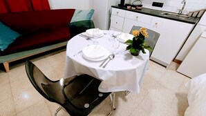 Zona de menjador amb una taula rodona preparada per a dues persones, una cadira, una planta petita en test i una cuina al fons.