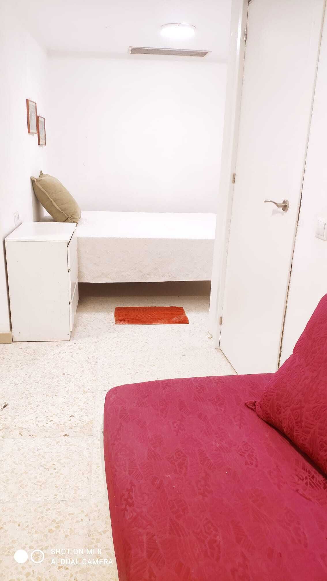 Una habitación pequeña y blanca con una cama, una mesita de noche, un tapete rojo y una puerta.