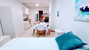 Un dormitori amb un llit, una taula i cadires, una cuineta i una escala.