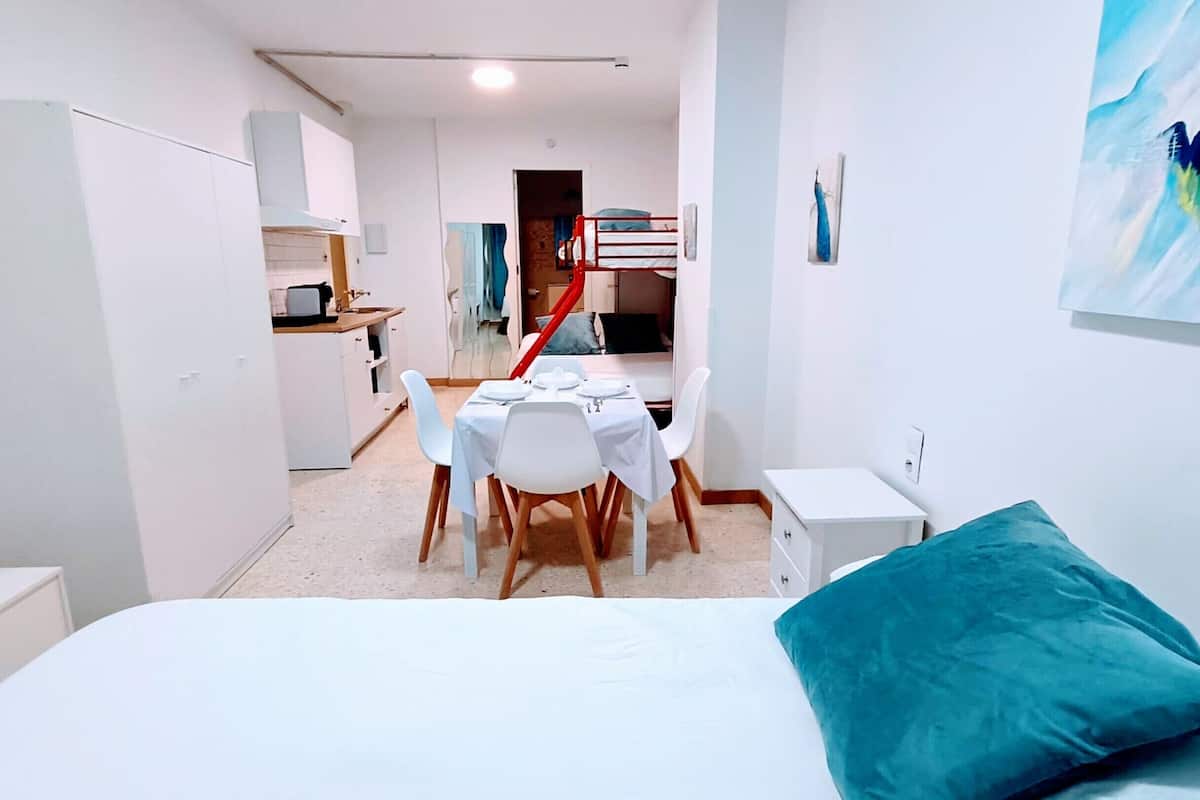A bedroom with a bed, a table with chairs, a kitchenette, and a staircase.