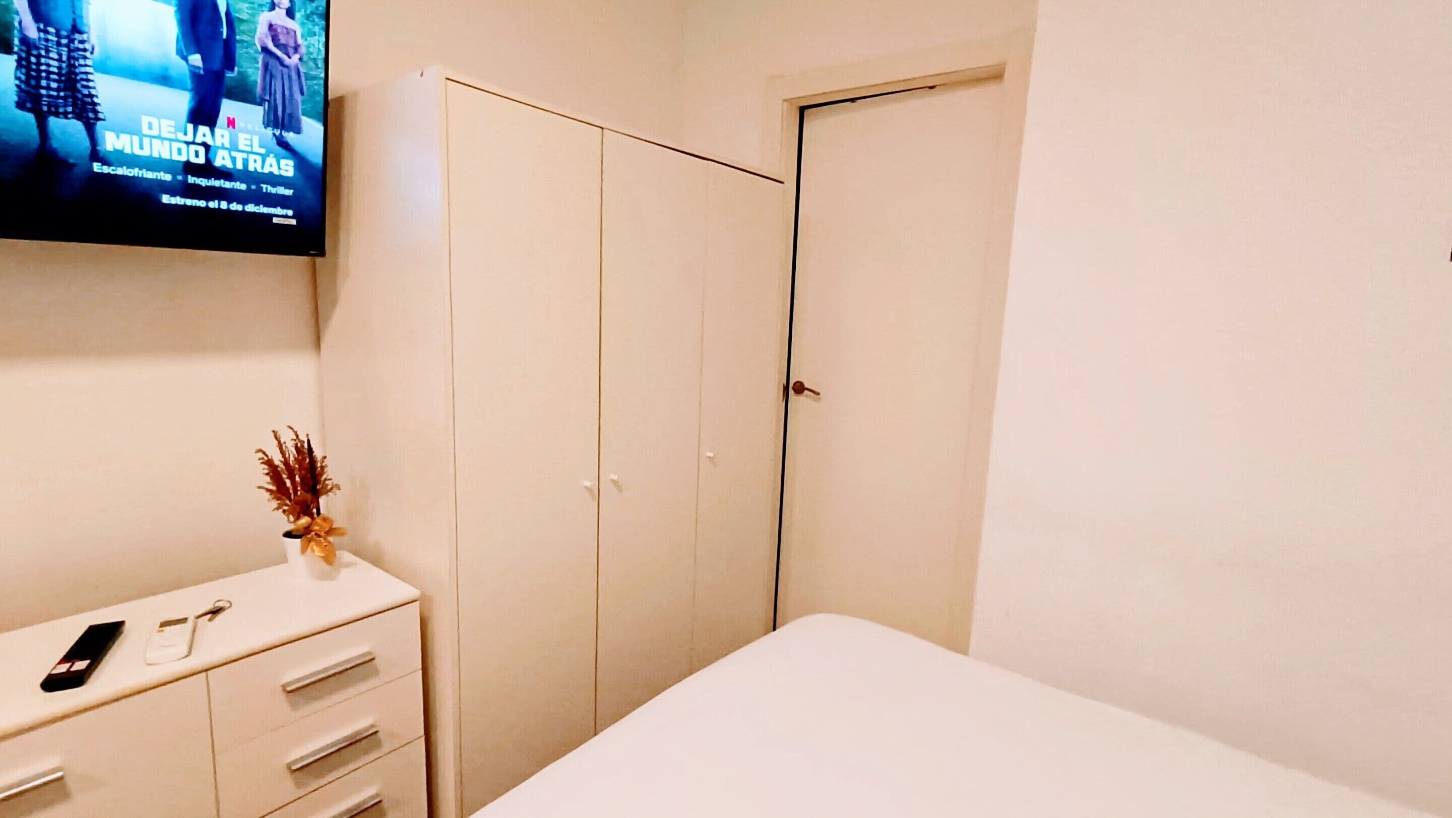 Un dormitorio con cama, cómoda con un florero y televisor mostrando un cartel de película.