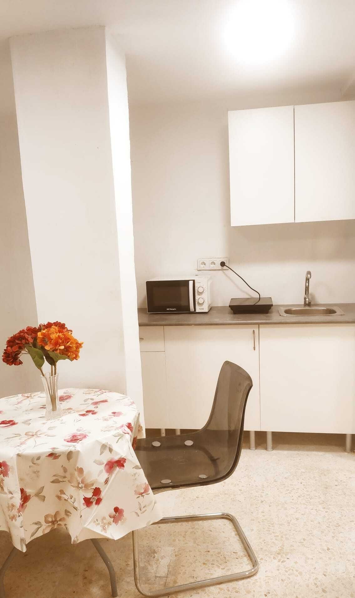 Una cocina pequeña y limpia con microondas, lavabo y armarios blancos. Hay una mesa con mantel floreado y un florero con flores.