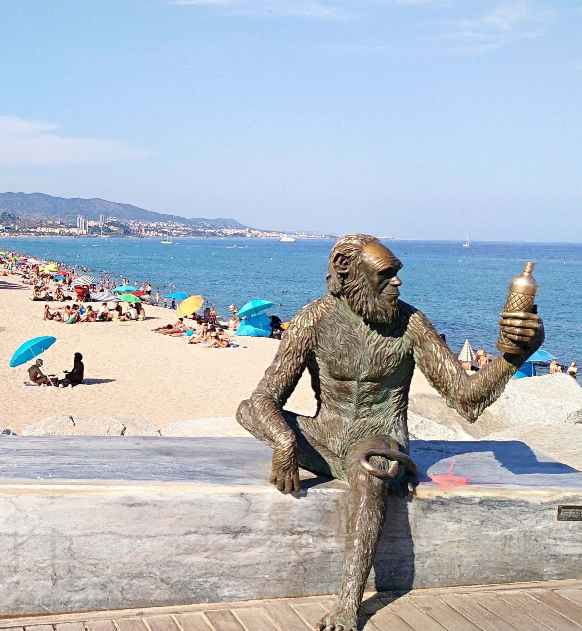 Una estatua de bronce de un hombre sosteniendo una botella sobre una pasarela de madera, con una playa y el océano al fondo.