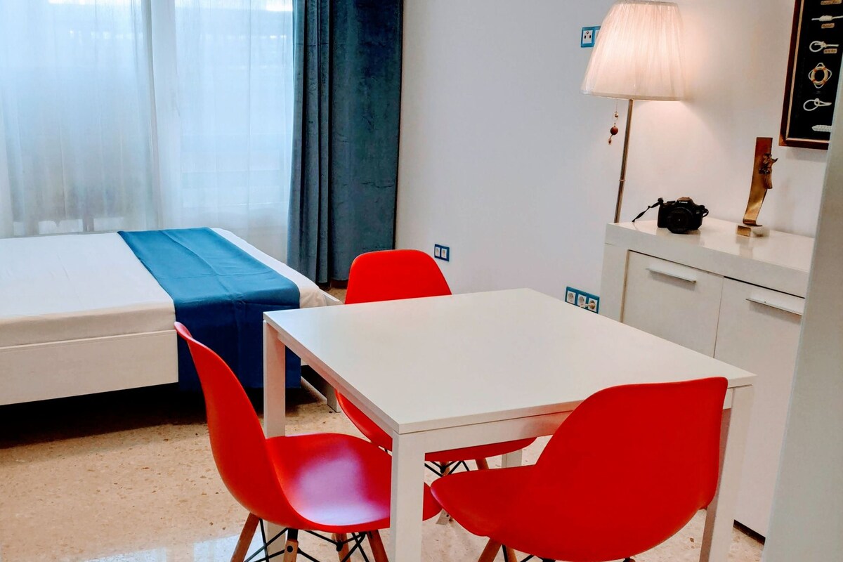 Una habitación de hotel pequeña con una cama, un sofá compacto, una mesa de comedor con sillas rojas, una lámpara y una cámara sobre la mesa.