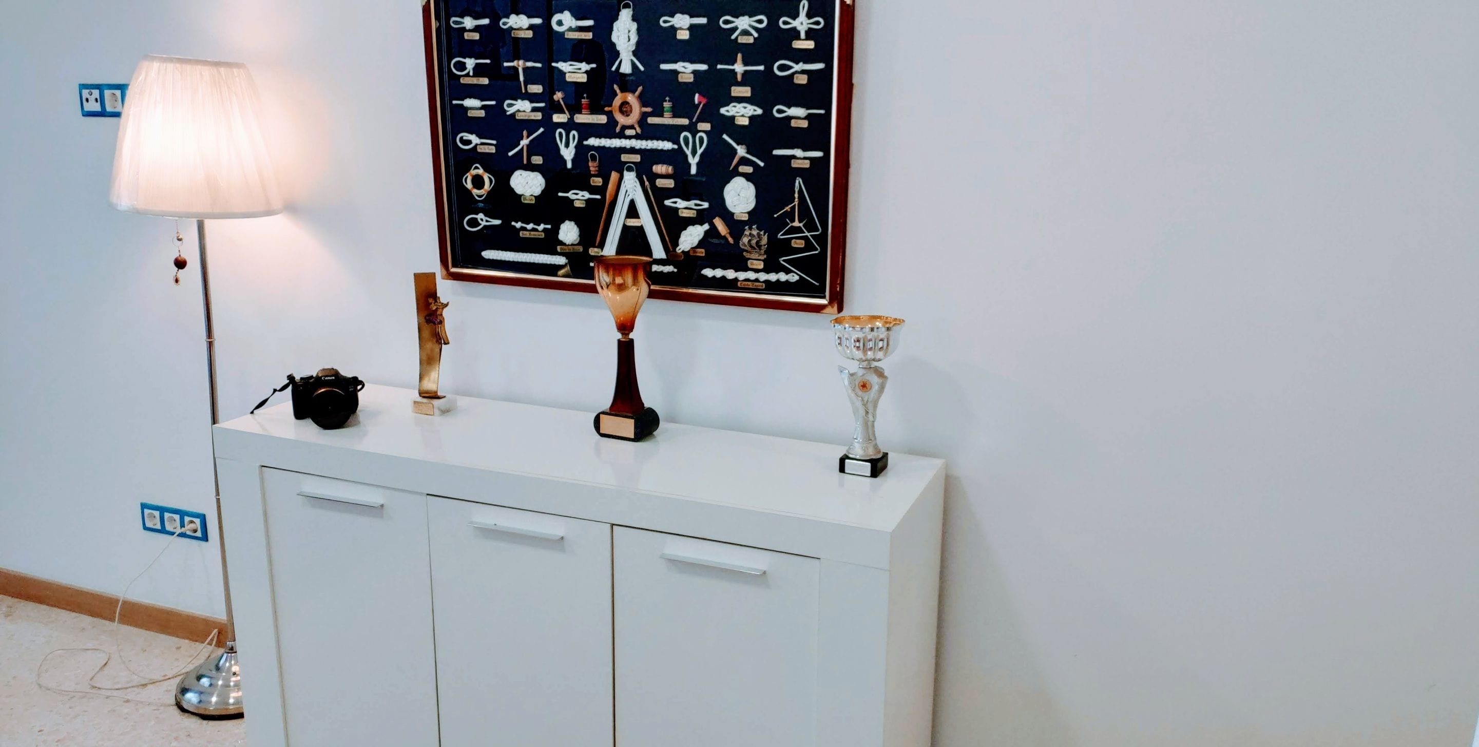 A white console table with a framed collection of knots and a camera on top.