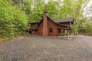 A log cabin with a chimney, a porch, and a gravel driveway surrounded by trees.