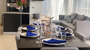 A dining area with a dark wooden table set for four, surrounded by grey upholstered chairs. A modern kitchen with a black cabinet and a flat-screen TV is visible in the background. - luxury Two Bedroom Private Home/ your ideal way to vacation (St george)