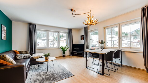 Een moderne woonkamer met een bank, een eettafel met stoelen, een kroonluchter en grote ramen met gordijnen.