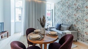 Ein modernes Wohnzimmer mit Essbereich, mit einem grauen Sofa, einem Holzesstisch gedeckt mit Tellern und Besteck sowie einem großen Fenster mit Vorhängen.