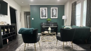 A living room with a dark wood floor, two black armchairs, a green sofa, a black piano, and two framed horse portraits on the wall.