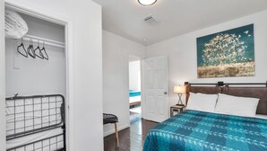 A bedroom with a bed, a nightstand, a lamp, a painting, a closet with hangers, and a chair.