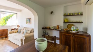 Een gezellige woonkamer met een houten eettafel, een beige bank en een grote groene schaal op de tafel. Er is een raam met uitzicht op groen.