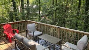 Una terraza de madera con mobiliario exterior, incluyendo una silla roja y sillas con cojines, rodeada de abundante follaje.