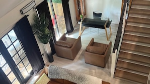 Amplia sala de estar con un sofá de cuero marrón, una mesa de centro redonda de madera y una mesa de comedor con sillas. Una escalera se ve a la derecha, que conduce a un piso superior.