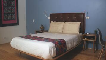 Uma cama bem arrumada com um edredom colorido, uma mesa de cabeceira com abajur e uma cadeira.