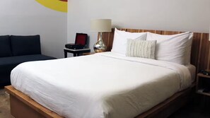 A hotel room with a bed, a bedside table with a lamp, a small table with a red case, and a dark-colored sofa. - Hotel Brecha (Tijuana)