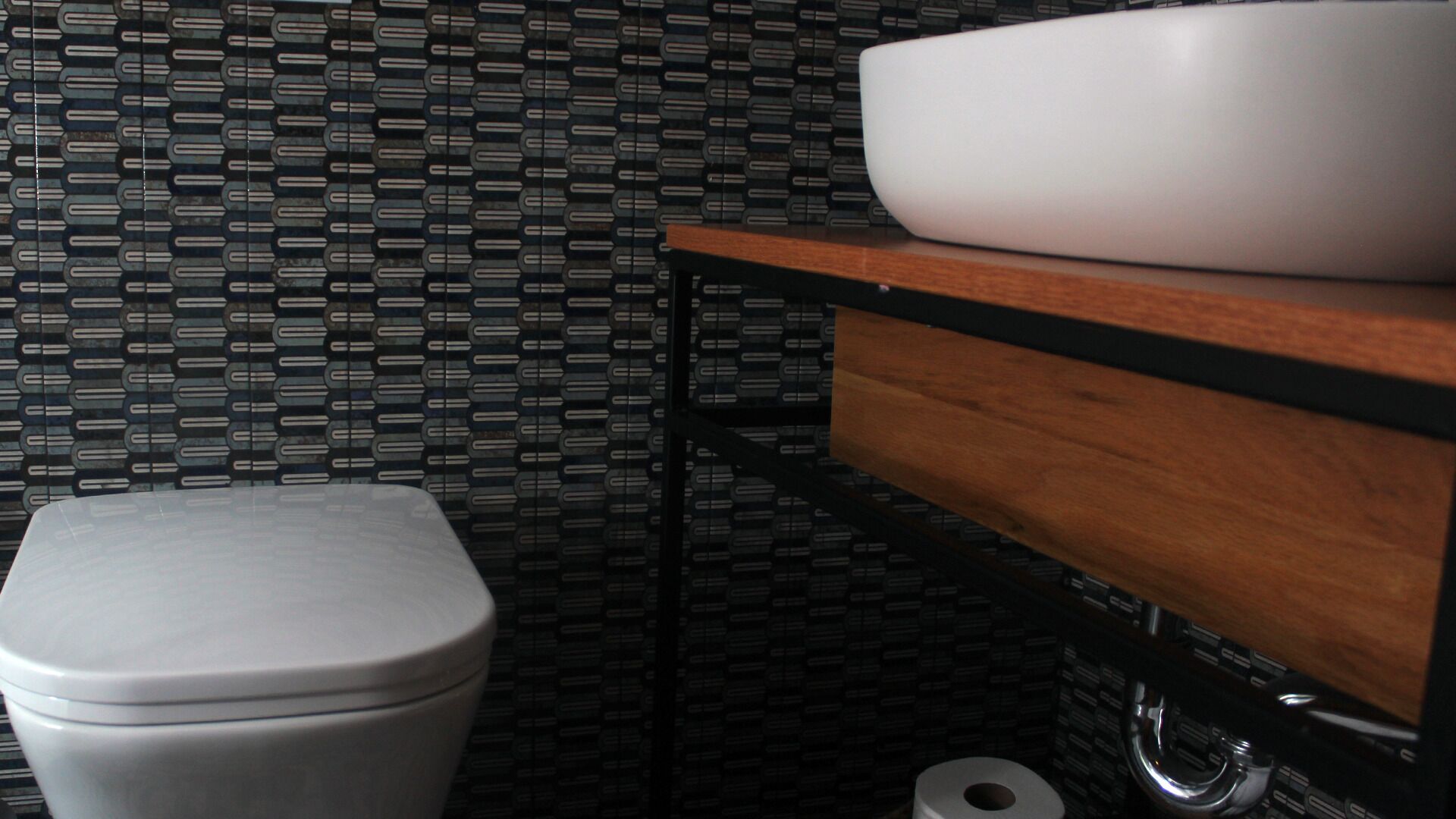 A bathroom with a toilet, a sink on a wooden shelf, and a patterned wall.