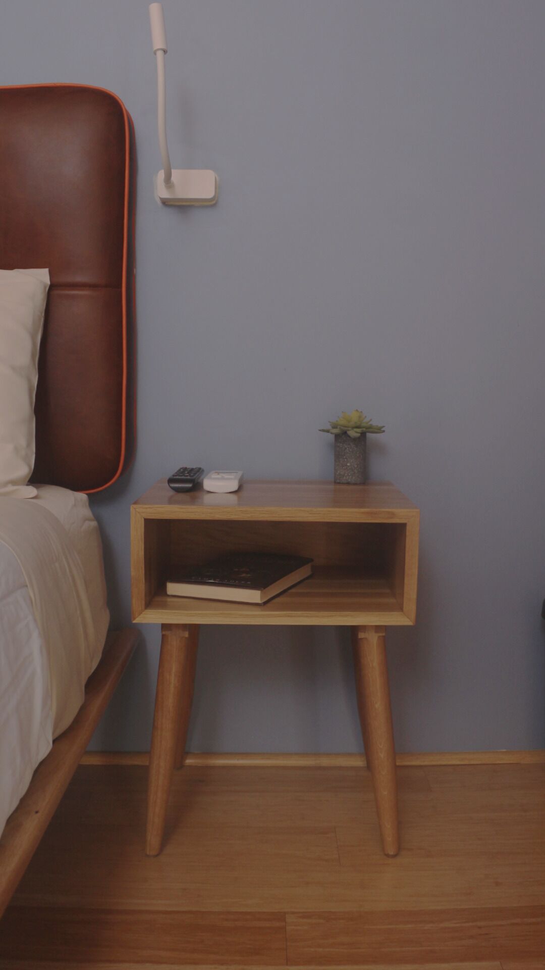 Un dormitorio con una mesita de noche de madera, una lámpara, un control remoto, un libro y una planta pequeña.