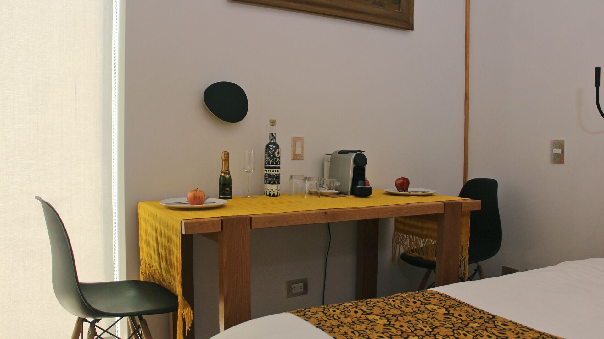 A hotel room with a wooden desk, a chair, a bed, and a small round table with a bottle, glasses, and an apple.