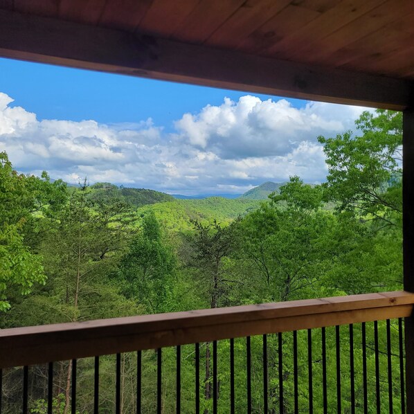 Une vue depuis un balcon en bois donnant sur un paysage verdoyant et luxuriant, avec des collines et un ciel partiellement nuageux.