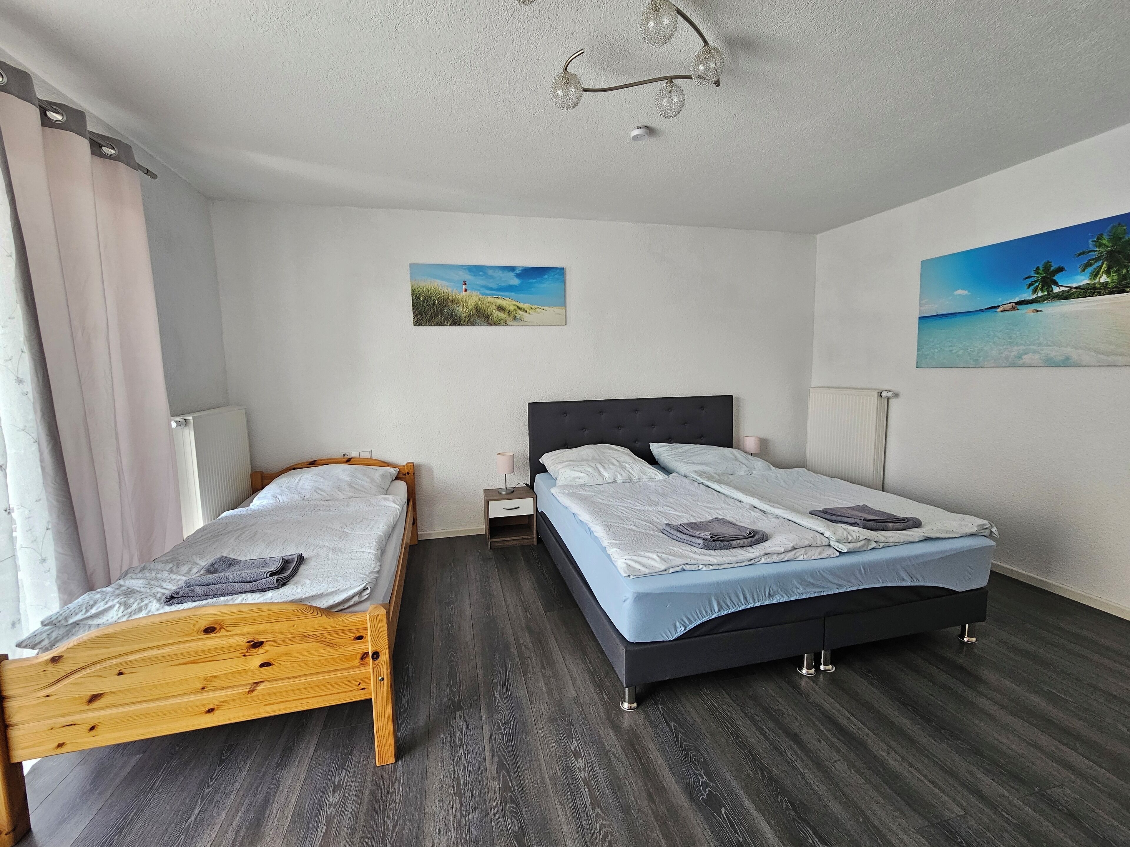 Un dormitorio con dos camas, una de madera a la izquierda y una tapizada de color negro a la derecha. Hay dos mesitas de noche, una con una lámpara y la otra con un cajón pequeño. El techo y las paredes son blancos, y hay dos cuadros en las paredes.