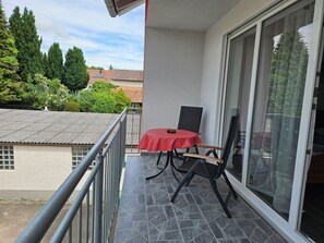 A balcony with a table and chairs, a sliding glass door, and a view of neighboring buildings and trees.