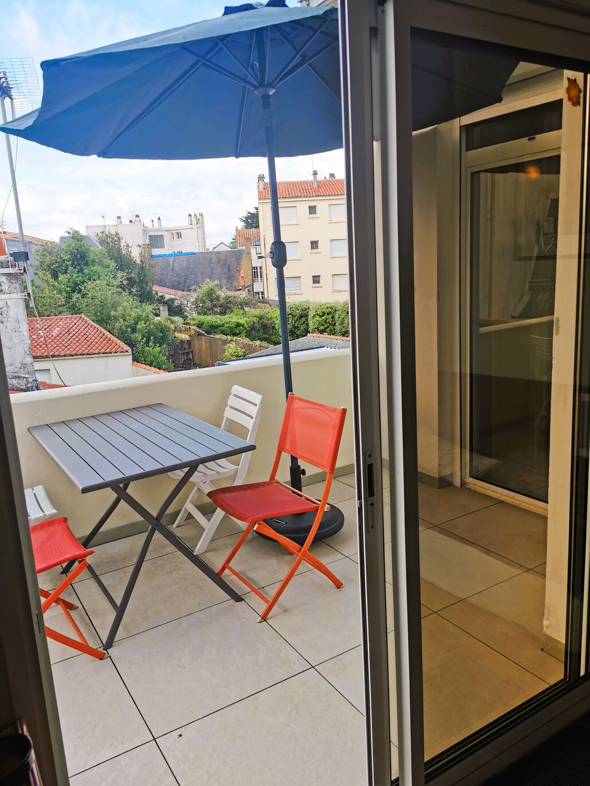 A balcony with a table and chairs, a blue umbrella, and a view of buildings in the background.
